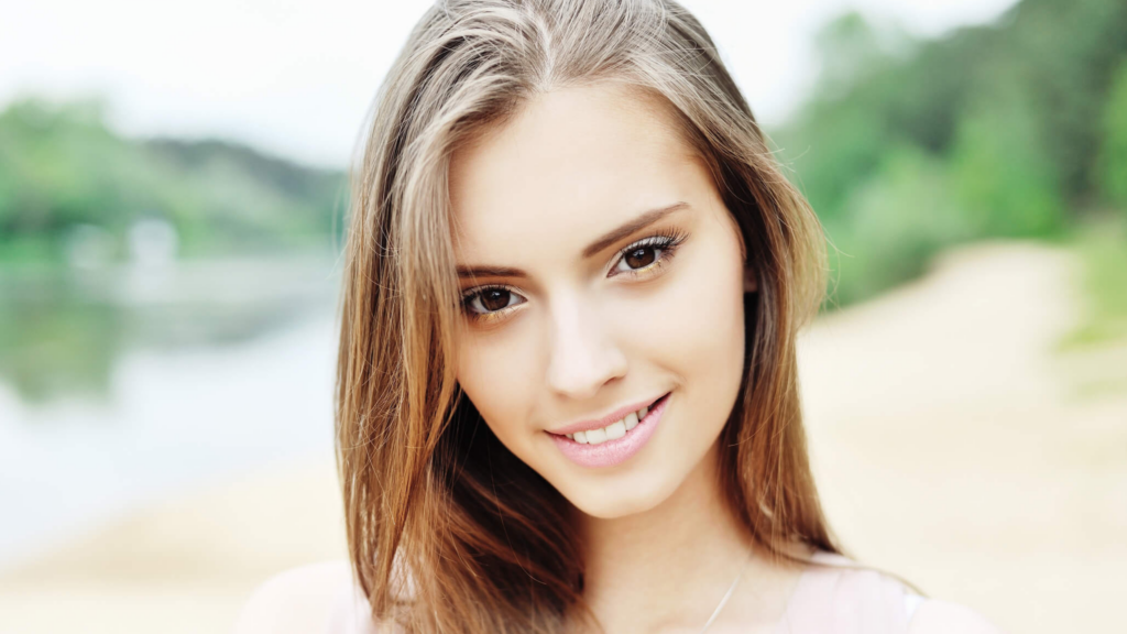 Close-up portrait of a smiling woman outdoors with a blurred green, sandy background behind her